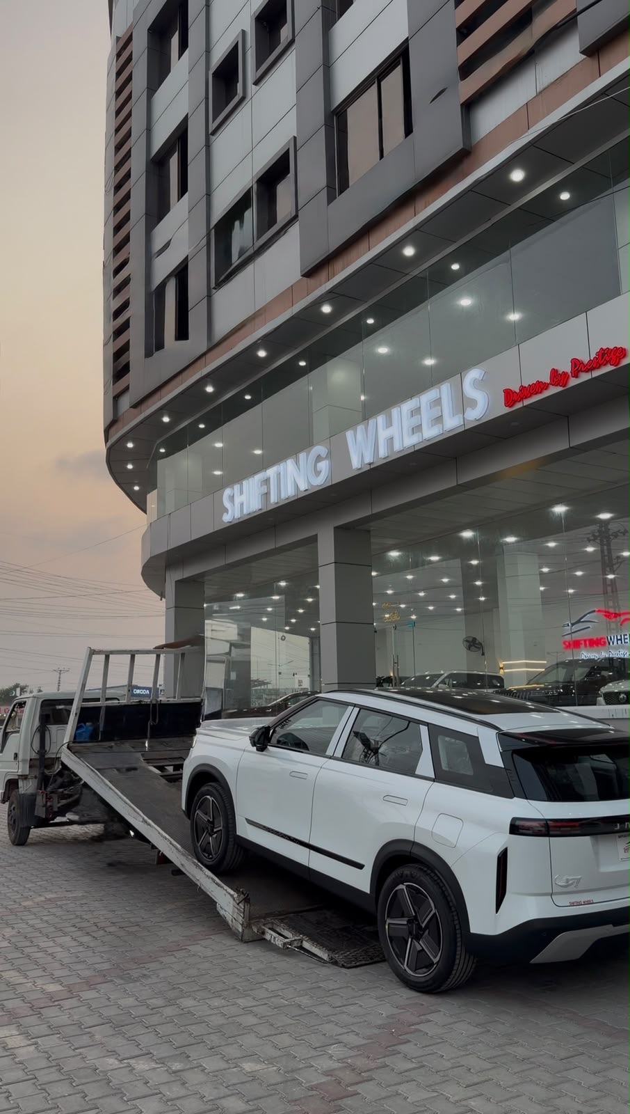 A white SUV being unloaded from a car carrier outside the Shifting Wheels showroom at dusk
